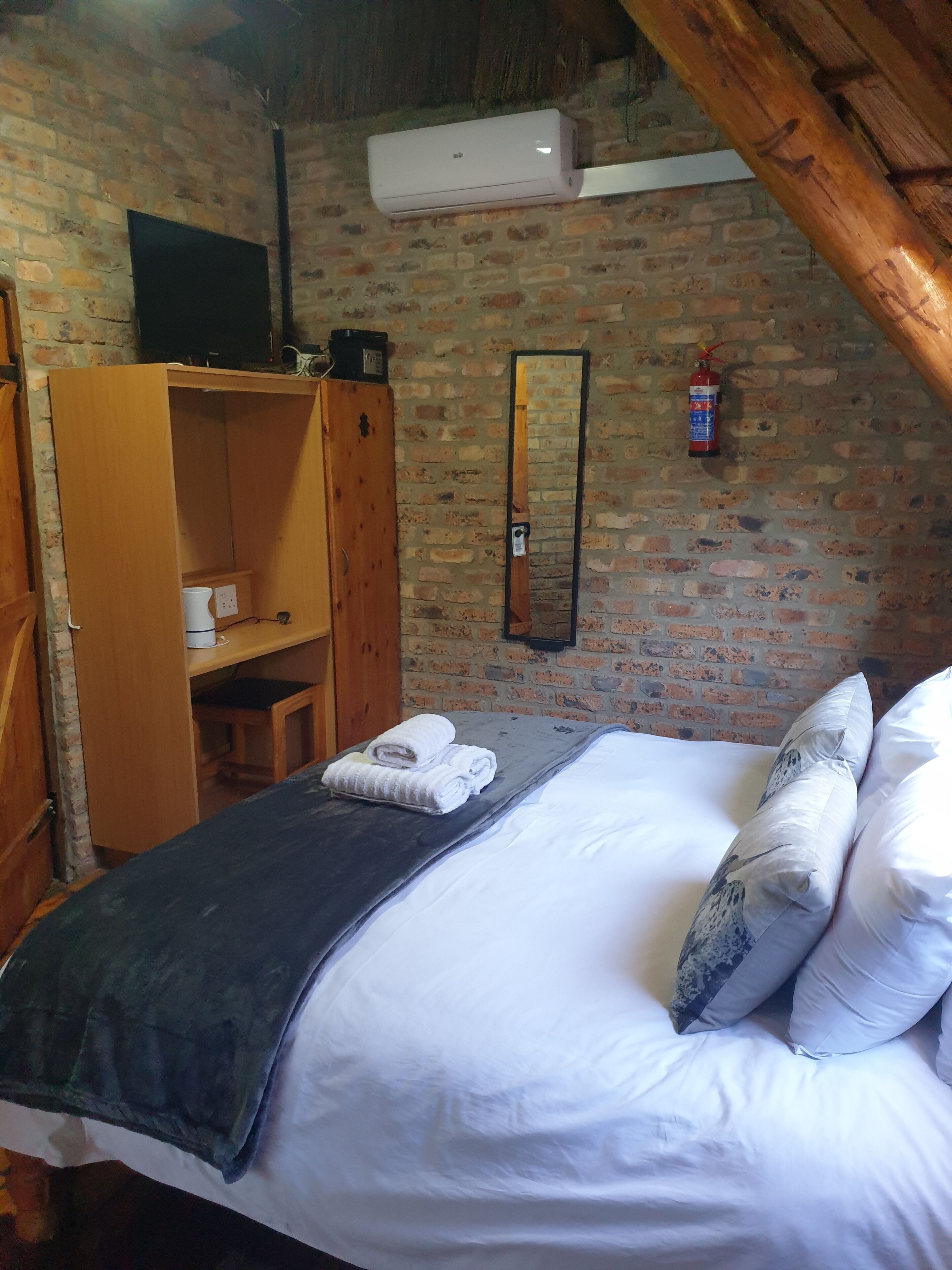 Un dormitorio con pared de ladrillo, armario de madera, cama con ropa blanca, televisor de pantalla plana y extintor.