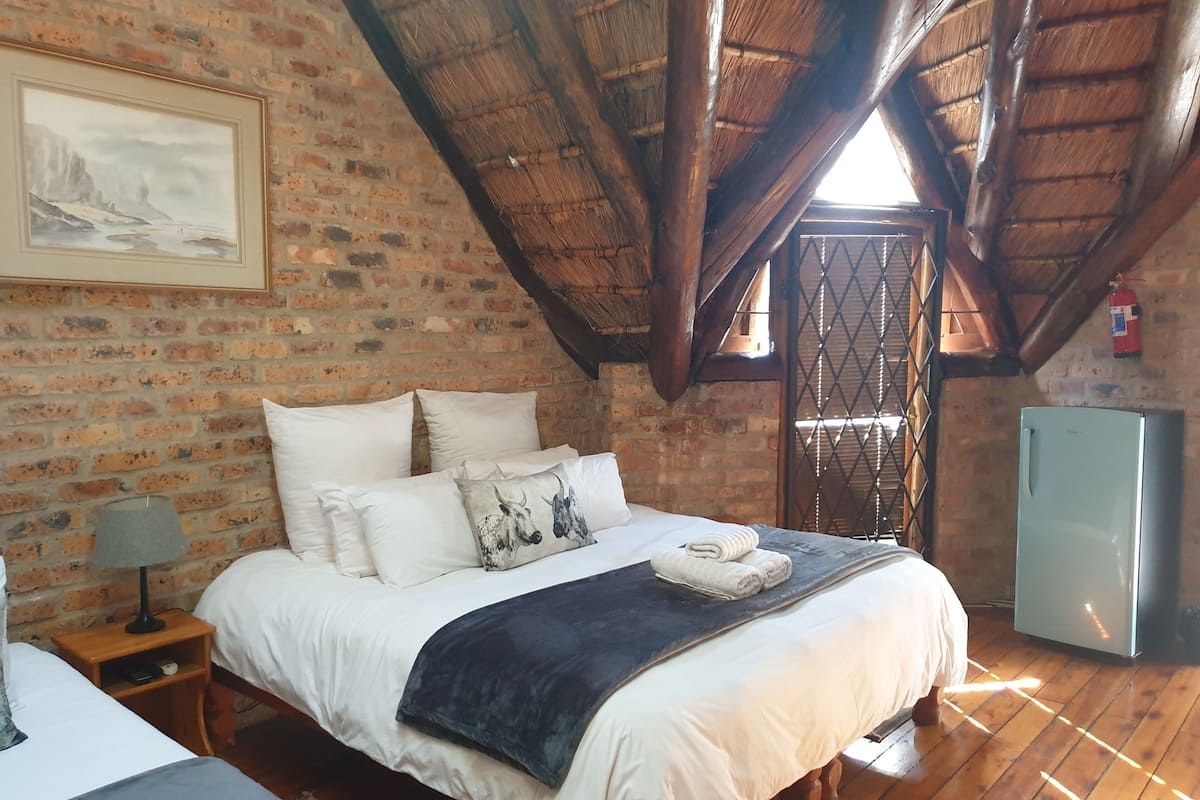 A bedroom with a brick wall, wooden beams, a bed with white bedding, a nightstand with a lamp, and a refrigerator.