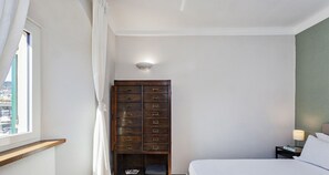 A bedroom with a bed, a nightstand, a lamp, a wooden dresser with multiple drawers, and a window with curtains.