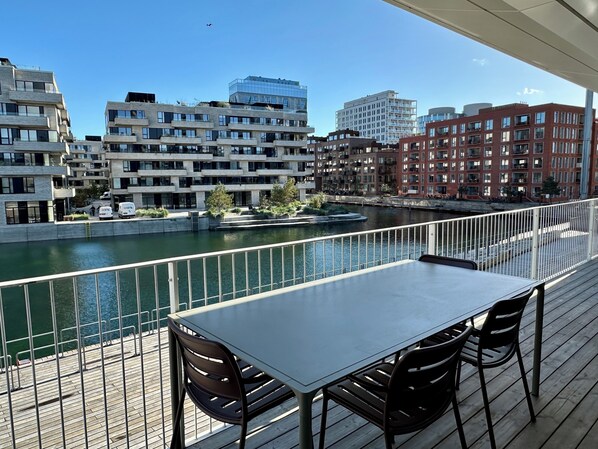 Un balcón con mesa y sillas que da a un río, con edificios modernos al fondo.