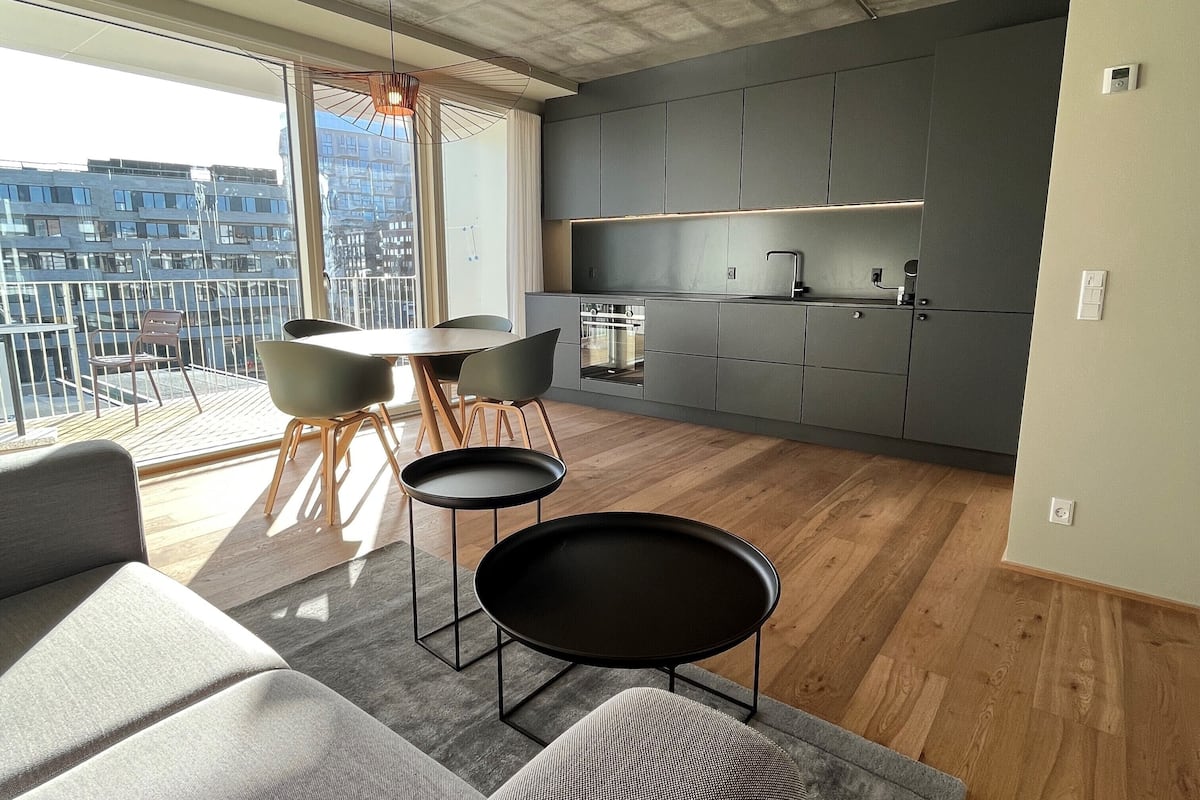 A modern kitchen with grey cabinets, a dining area with a round table and chairs, and a balcony with a view of buildings.