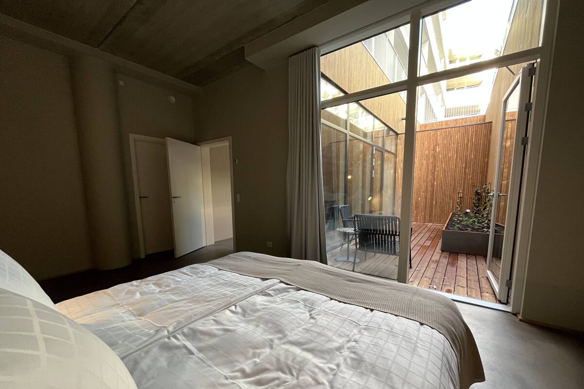 A bedroom with a large bed, a sliding glass door leading to a balcony with a wooden deck and a small table.