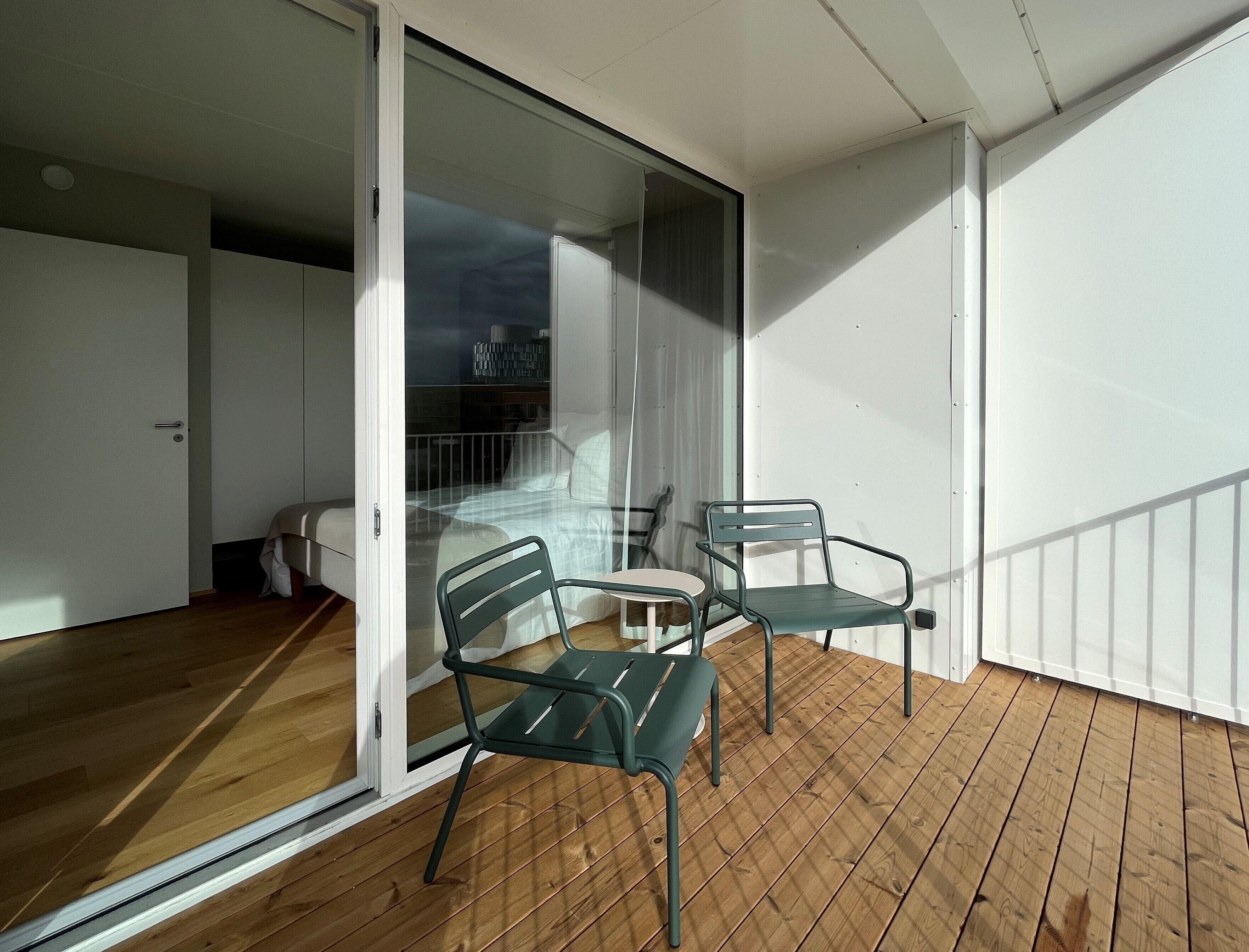 A balcony with wooden flooring, two green chairs, and a glass door leading to a bedroom.