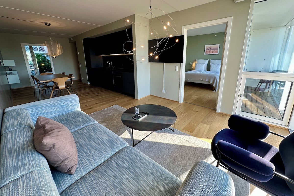 A modern living room with a sofa, a round coffee table, and a TV. There is a dining area with a table and chairs, and a kitchen in the background.