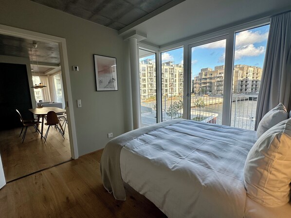 Un dormitorio con una cama grande, una zona de comedor con sillas y vistas a edificios a través de amplias ventanas.