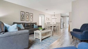 A spacious living room with a grey sofa, a blue armchair, and a coffee table. There is a dining area with a chandelier and a kitchen in the background. - Rockwall Duo - Cottage + Lodge Combo (Branson)