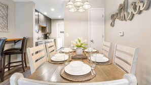 A dining area with a wooden table set for six, surrounded by white chairs, a chandelier, and a decorative wall piece. - Rockwall Duo - Cottage + Lodge Combo (Branson)