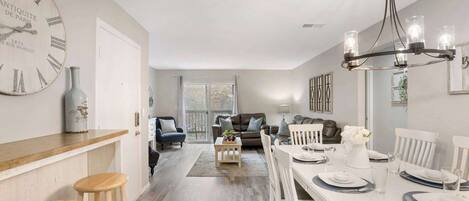 A spacious living area with a dining table set for four, a large wall clock, and a chandelier.
