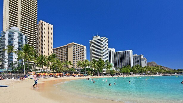 Una playa con agua turquesa cristalina, palmeras y un paisaje urbano con edificios altos al fondo.