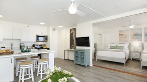 Una cocina moderna con armarios blancos, un microondas y un comedor con taburetes. Hay una cama con cabecera blanca y un televisor montado en la pared.
