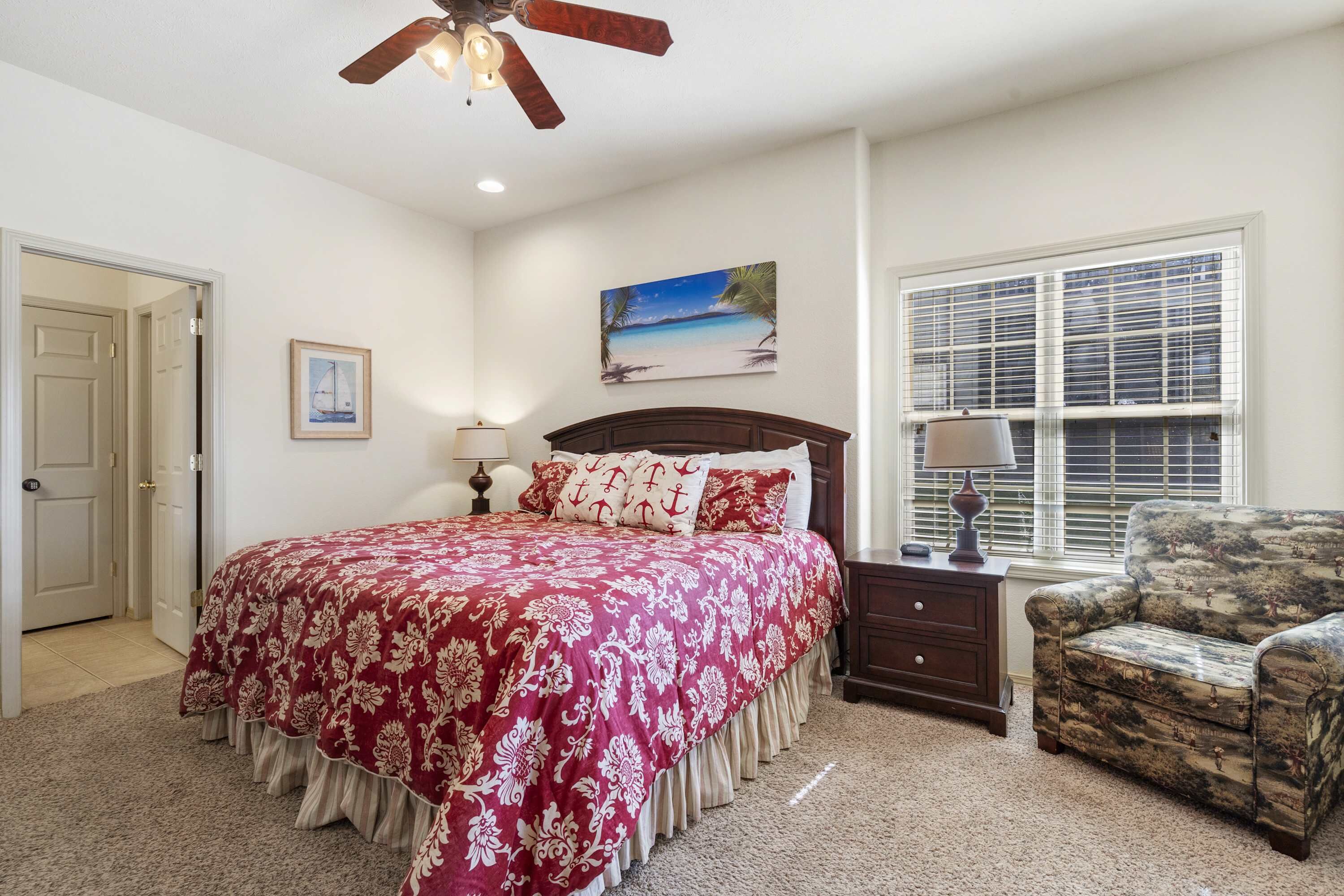 A bedroom with a bed, a nightstand, a chair, a ceiling fan, a painting, and a window with blinds.