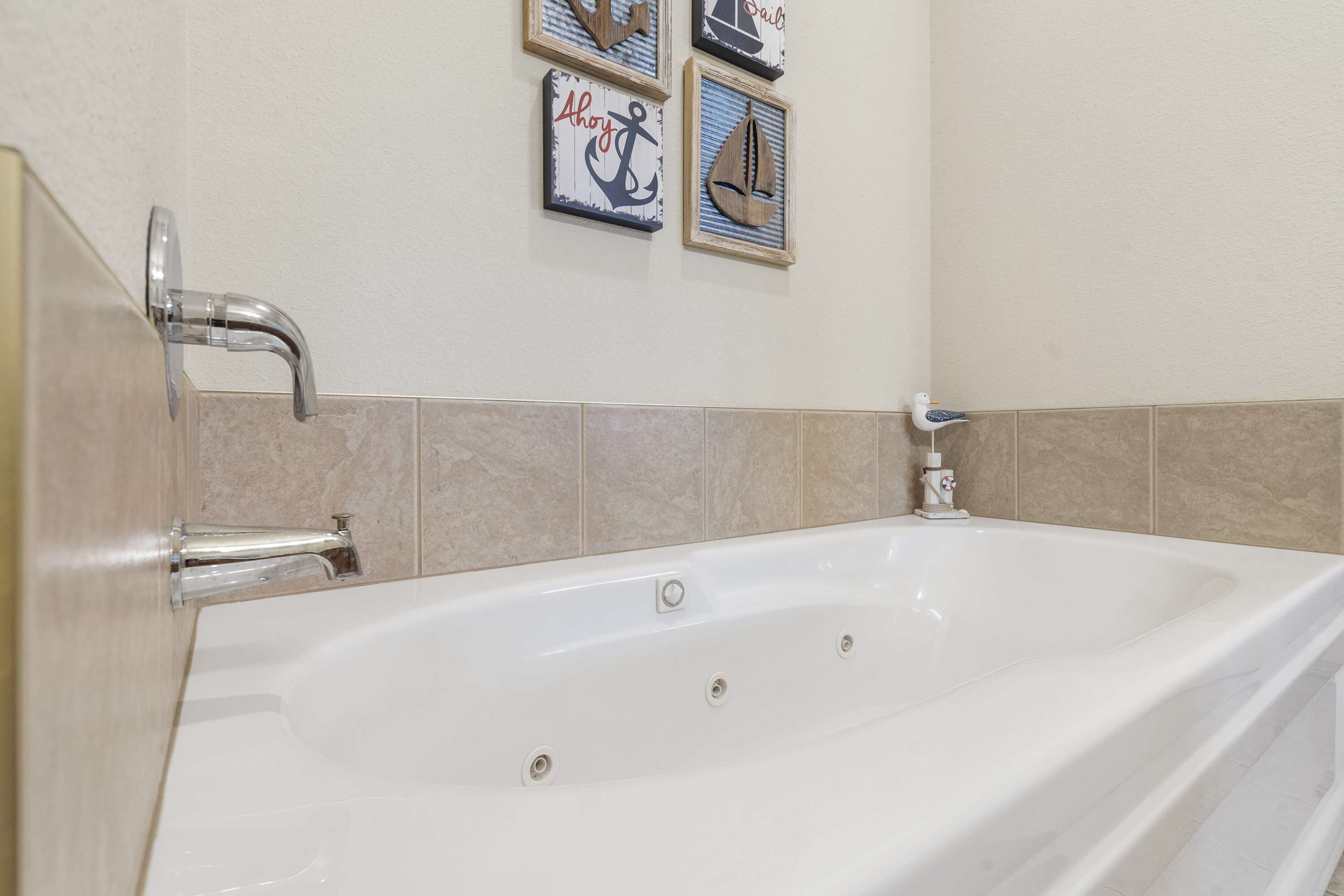 A white bathtub with a chrome faucet, beige tiled walls, and nautical-themed wall art.