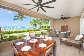 A dining area with a wooden table set for four, a ceiling fan, wicker furniture, and a view of the beach and ocean.