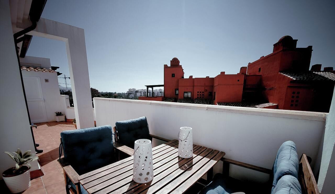 A rooftop terrace with a wooden table, two chairs, and a sofa. There are two white decorative vases on the table. The terrace has a view of red buildings in the background.