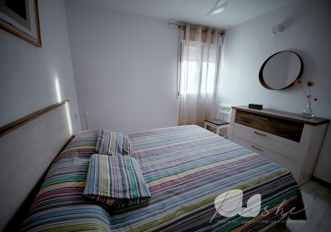 A bedroom with a bed, a bedside table, a dresser, a mirror, and a window with curtains.