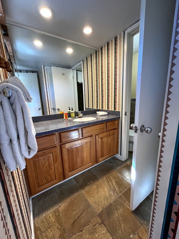 A bathroom with a wooden vanity, a sink, and a mirror. There are towels hanging on a rack, and a shower is visible in the background.