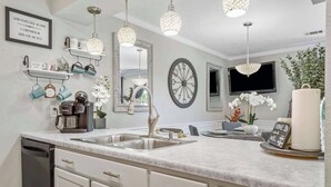 Uma cozinha moderna com armários brancos, uma pia dupla e iluminação pendente. Há objetos decorativos nas prateleiras e um espelho grande acima da pia.