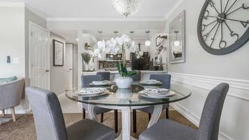 A dining area with a glass table, grey upholstered chairs, a white vase with orchids, and a large wall clock.
