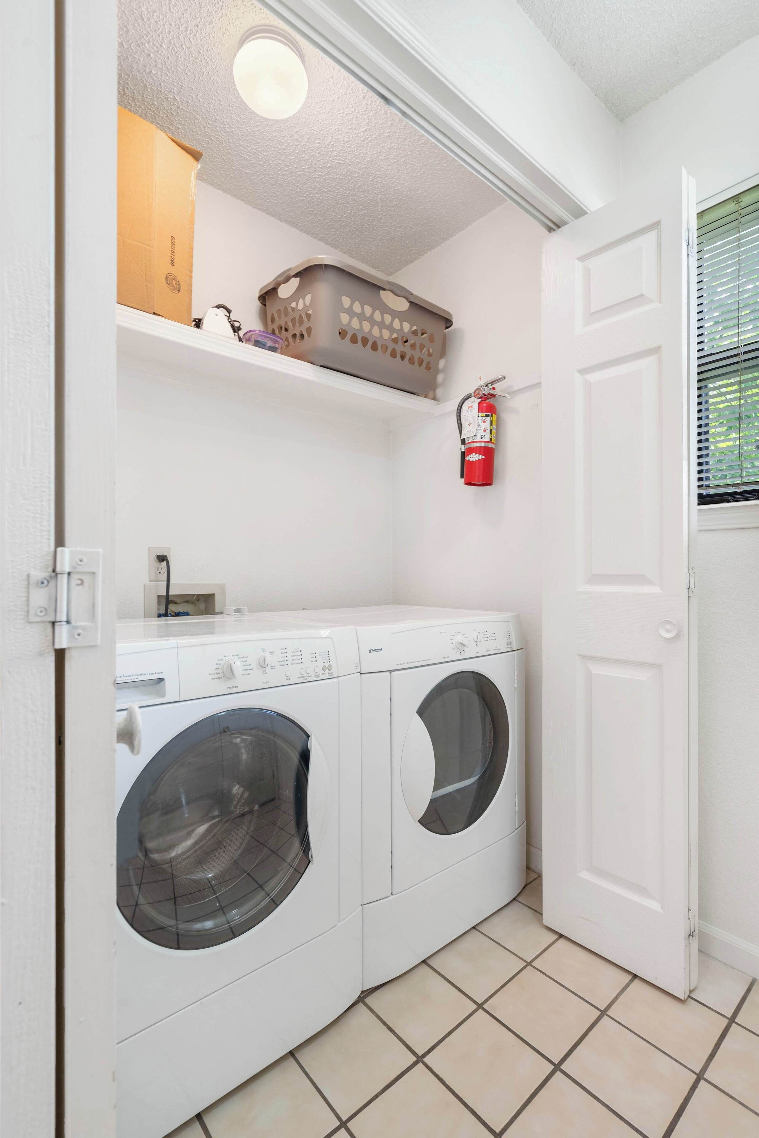 A laundry room with two front-loading washing machines, a shelf with a basket, a fire extinguisher, and a door.