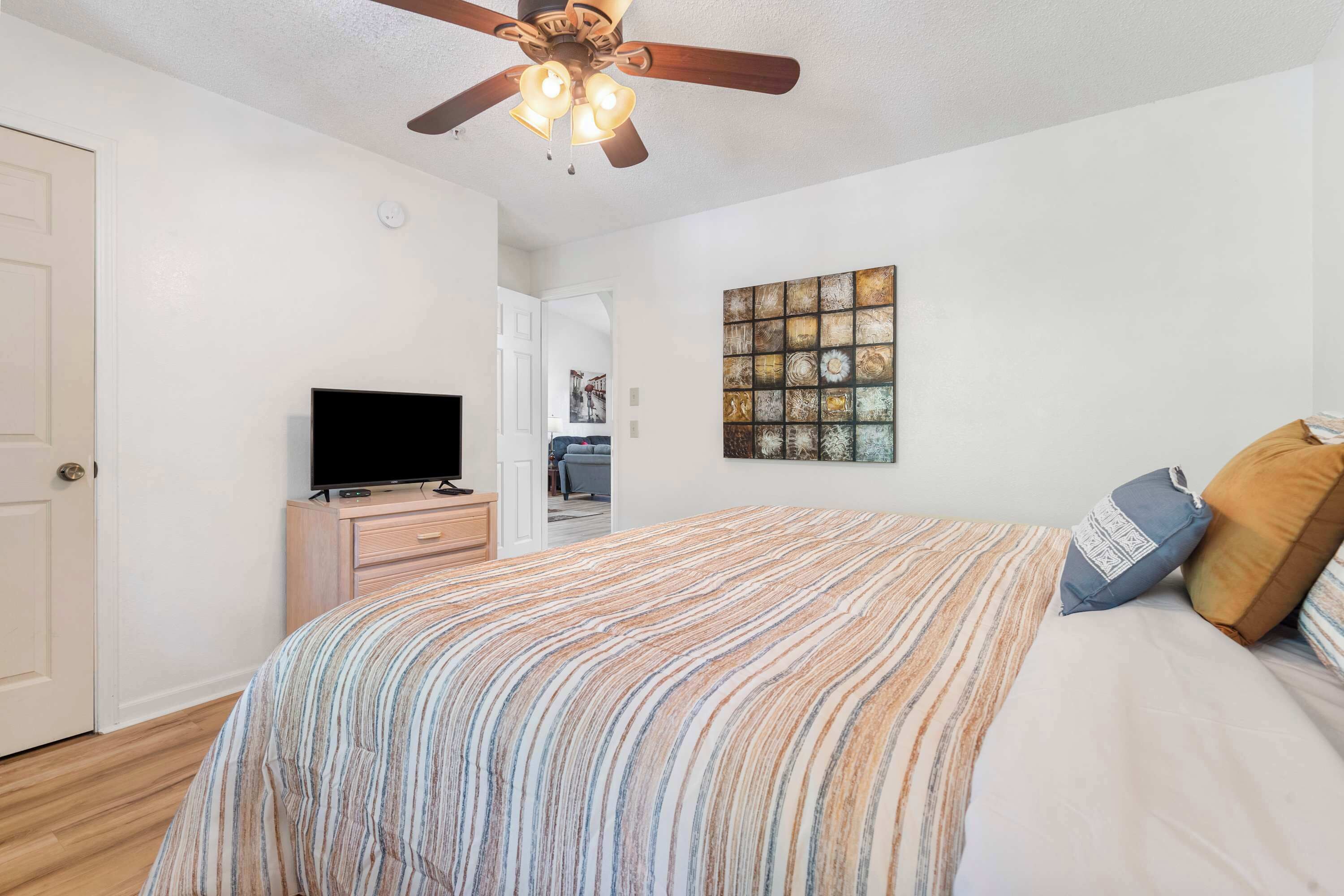 A bedroom with a bed, a television on a wooden dresser, a ceiling fan, and a wall art piece.