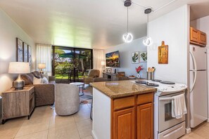 A modern kitchen with a central island, built-in oven, and refrigerator. There is a dining area with a table and chairs, and a living area with a sofa and a lamp.