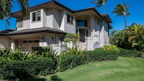 A two-story house with a garage, surrounded by lush greenery and palm trees.