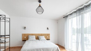 Un dormitorio con cama, cabecero de madera, dos mesitas de noche, lámpara colgante y ventana con cortinas de gasa.