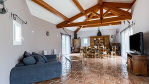 Un salón amplio con techo alto, vigas de madera a la vista, un sofá azul, una zona de comedor con muebles de madera y un televisor a la derecha.