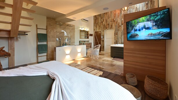 A modern hotel room with a large bed, a TV on the wall, a bathroom with a shower, and a view of a waterfall on the screen. - Instinct - Jacuzzi, Sauna, Apéritif dînatoire et petit déjeuner (Rocquigny)