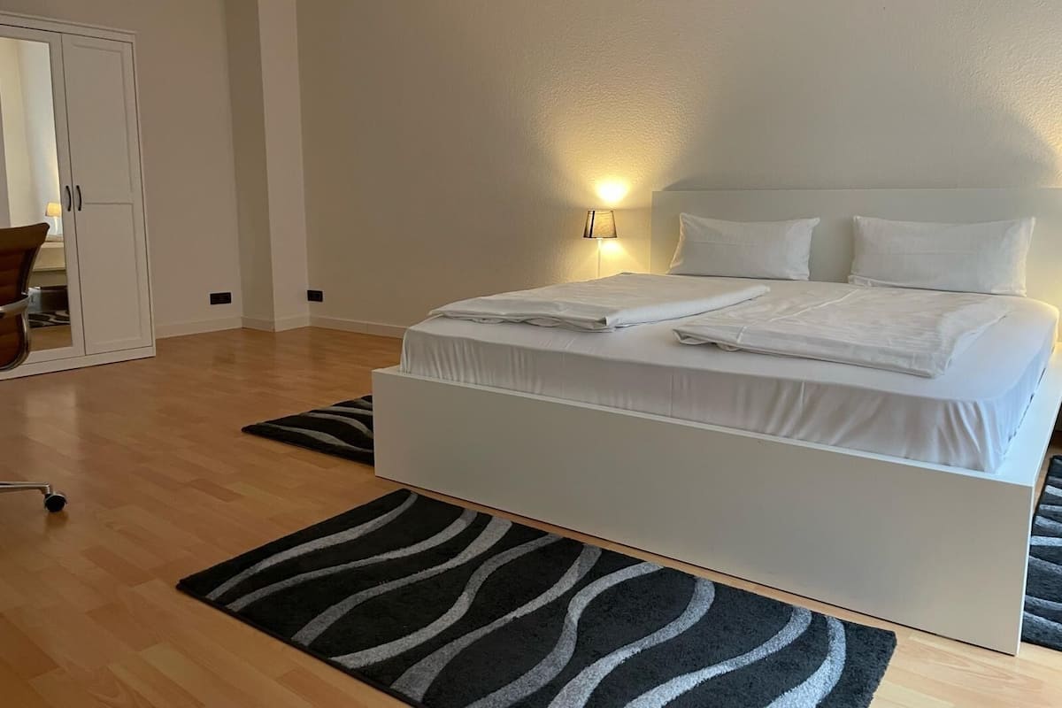 Ein ordentlich bezogenes Bett mit weißen Leinen in einem modernen Zimmer mit Holzboden und einem schwarz-weißen Teppich.