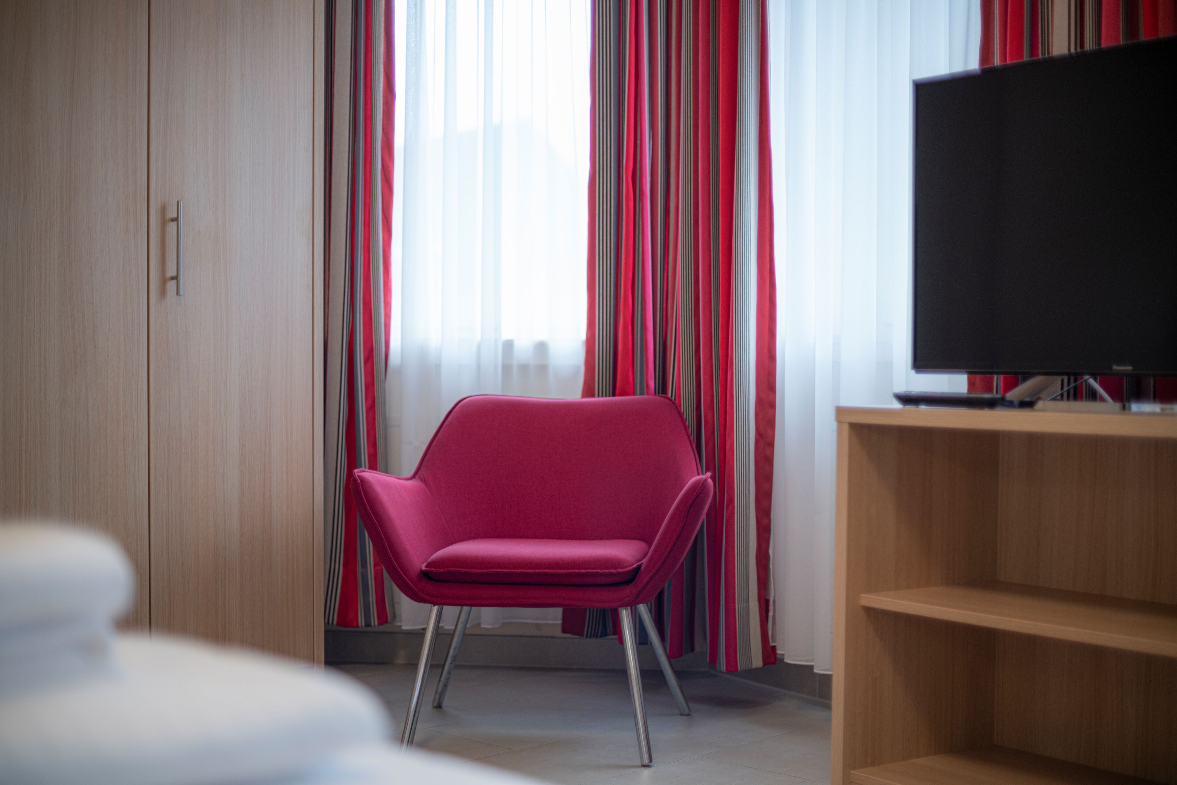 Une pièce avec un fauteuil rouge, un téléviseur posé sur un meuble en bois et une fenêtre ornée de rideaux à rayures.