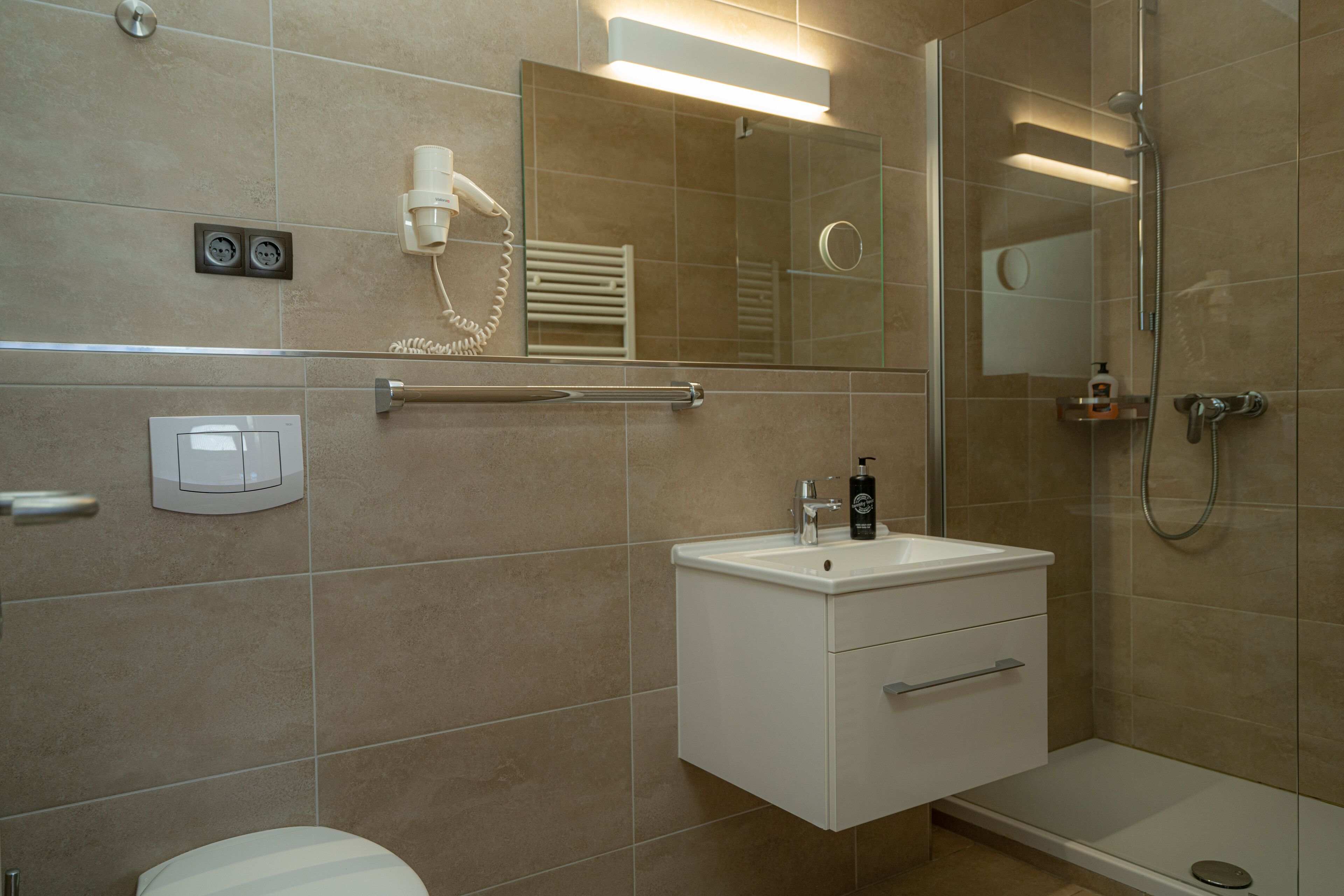 A modern bathroom with a toilet, sink, and shower. It includes a towel rack, a mirror, and a soap dispenser.