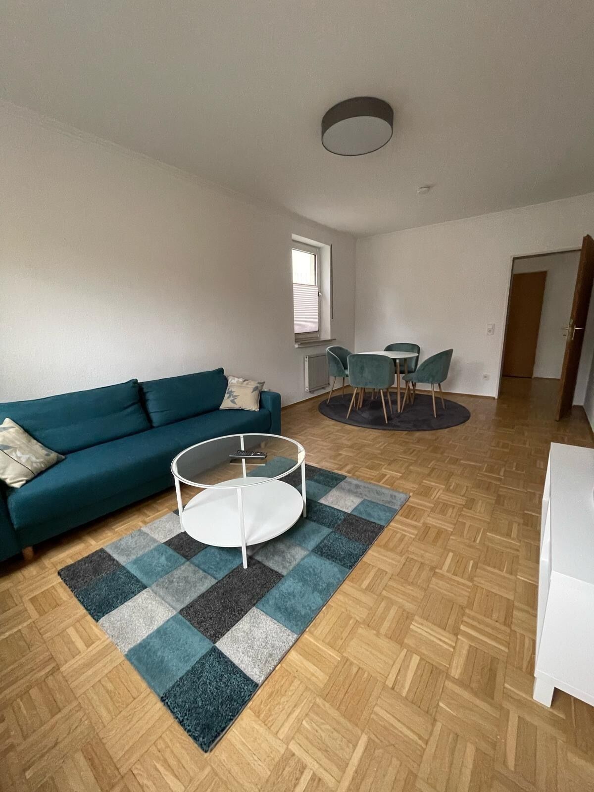 A living room with a teal sofa, a glass coffee table, and a dining area with a round table and chairs.