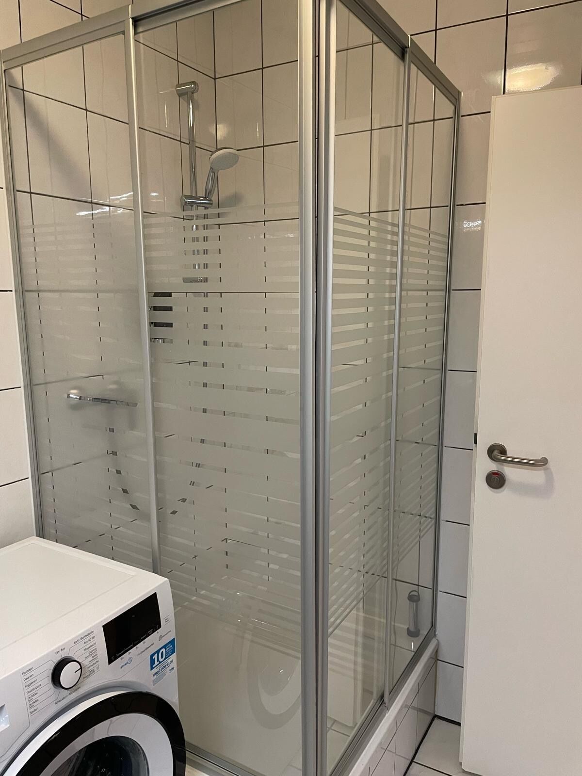 Un baño con ducha separada por vidrio, lavadora blanca y una puerta con picaporte.
