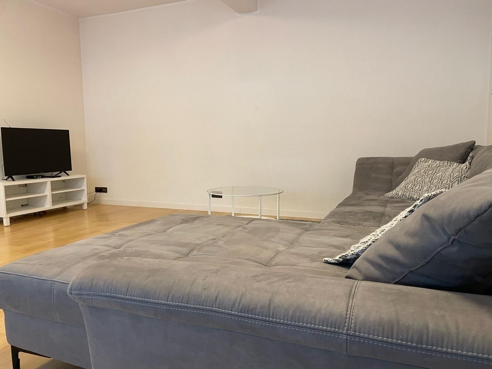 A living room with a grey sofa, a glass coffee table, a flat-screen TV on a white stand, and a patterned throw pillow.