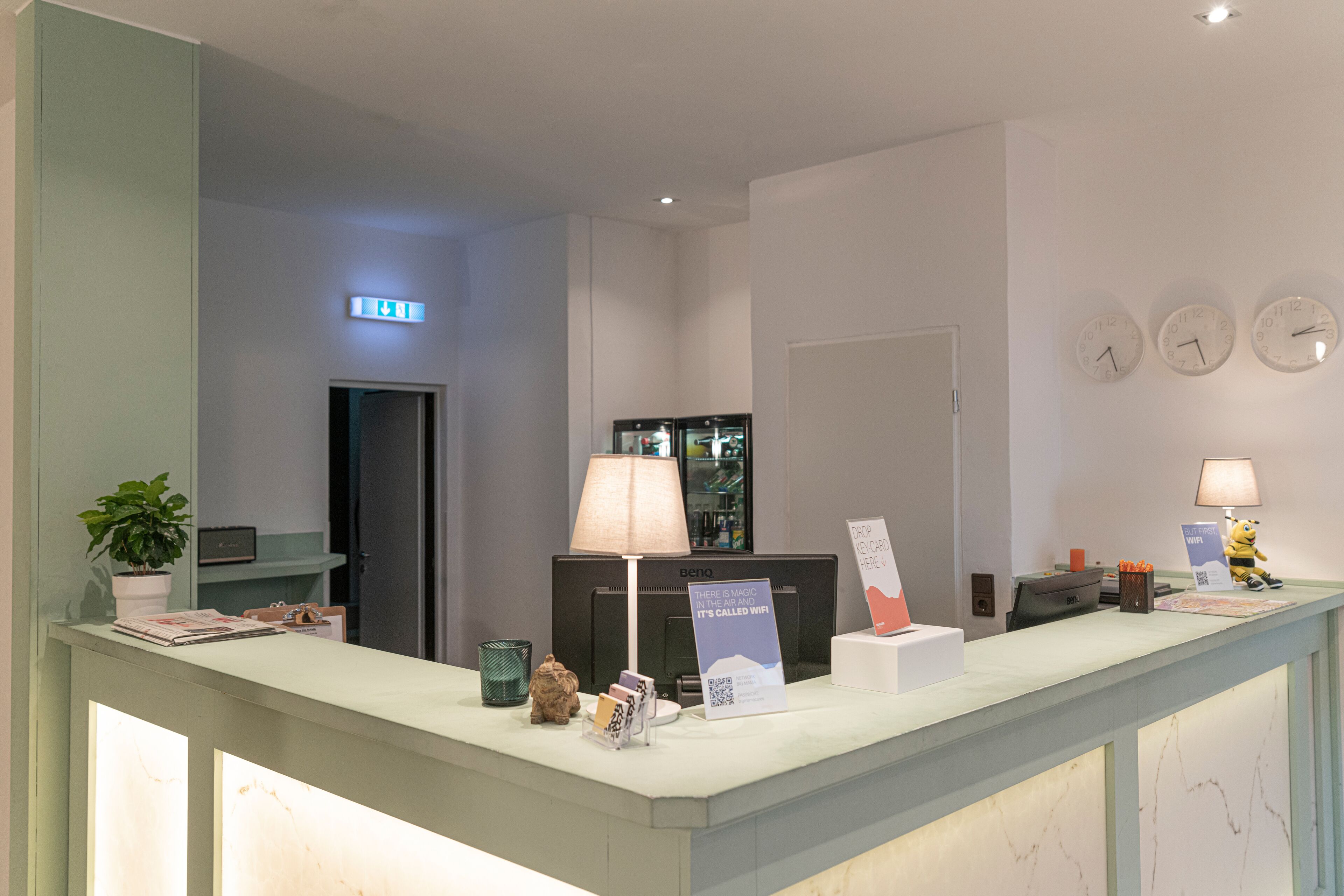 Een hotel receptiebalie met een computer, een lamp, een bord en een klok aan de muur.