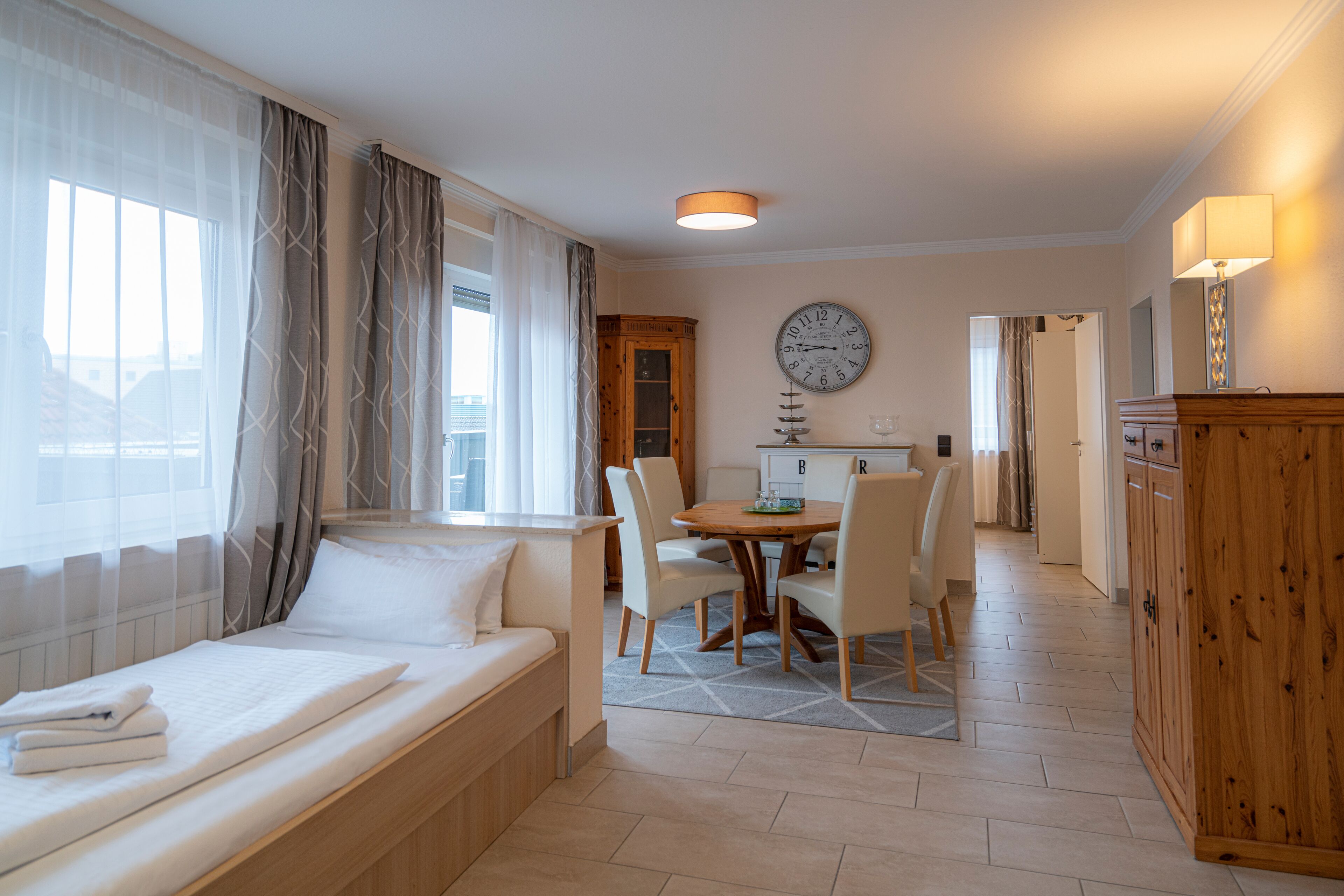 Quarto de hotel com cama, mesa de jantar com cadeiras, aparador de madeira e relógio de parede.