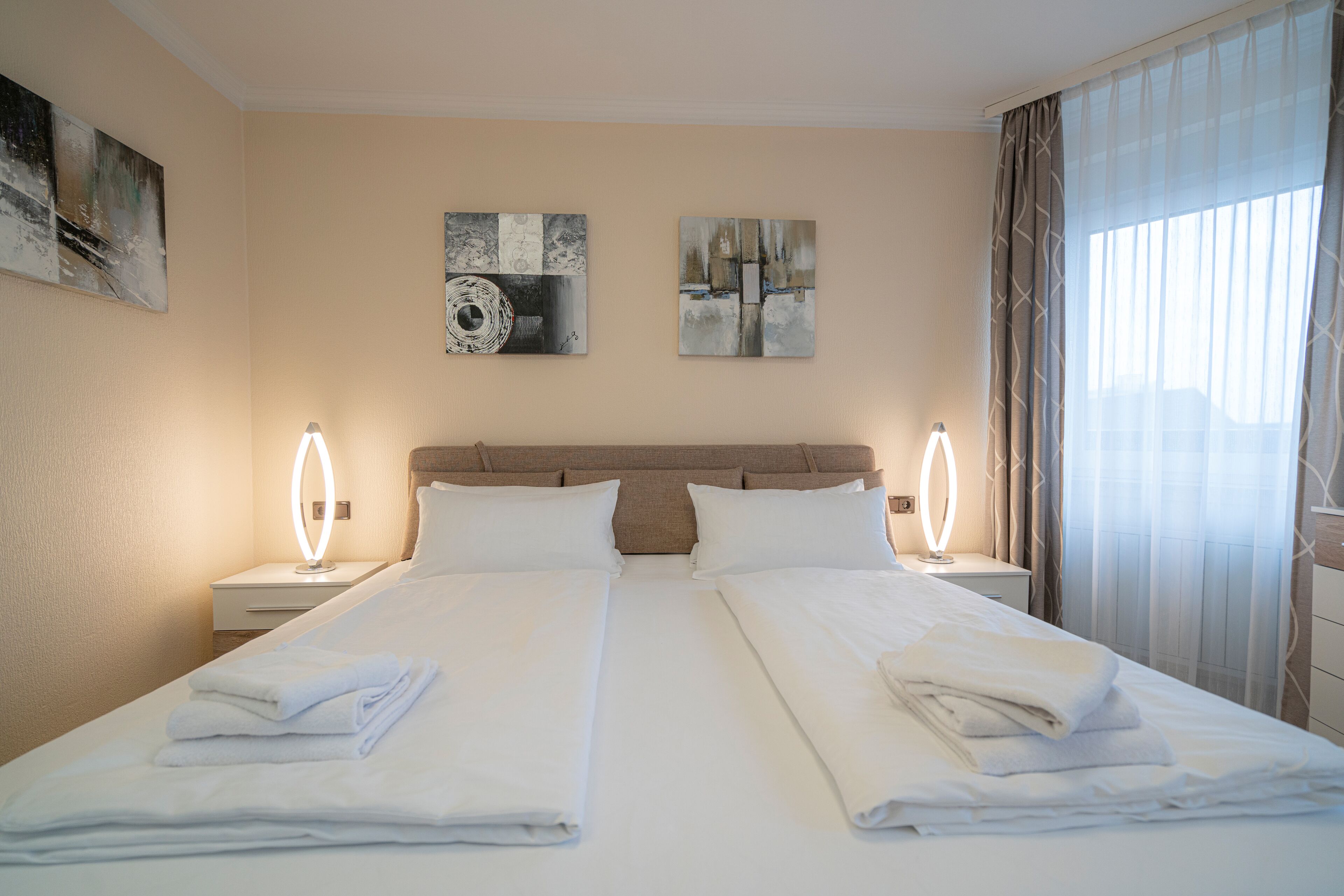 Habitación de hotel con dos camas, cabeceras, mesitas de noche con lámparas y obras de arte en las paredes.