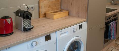 Cocina compacta con lavadora, cafetera y tabla de cortar de madera.