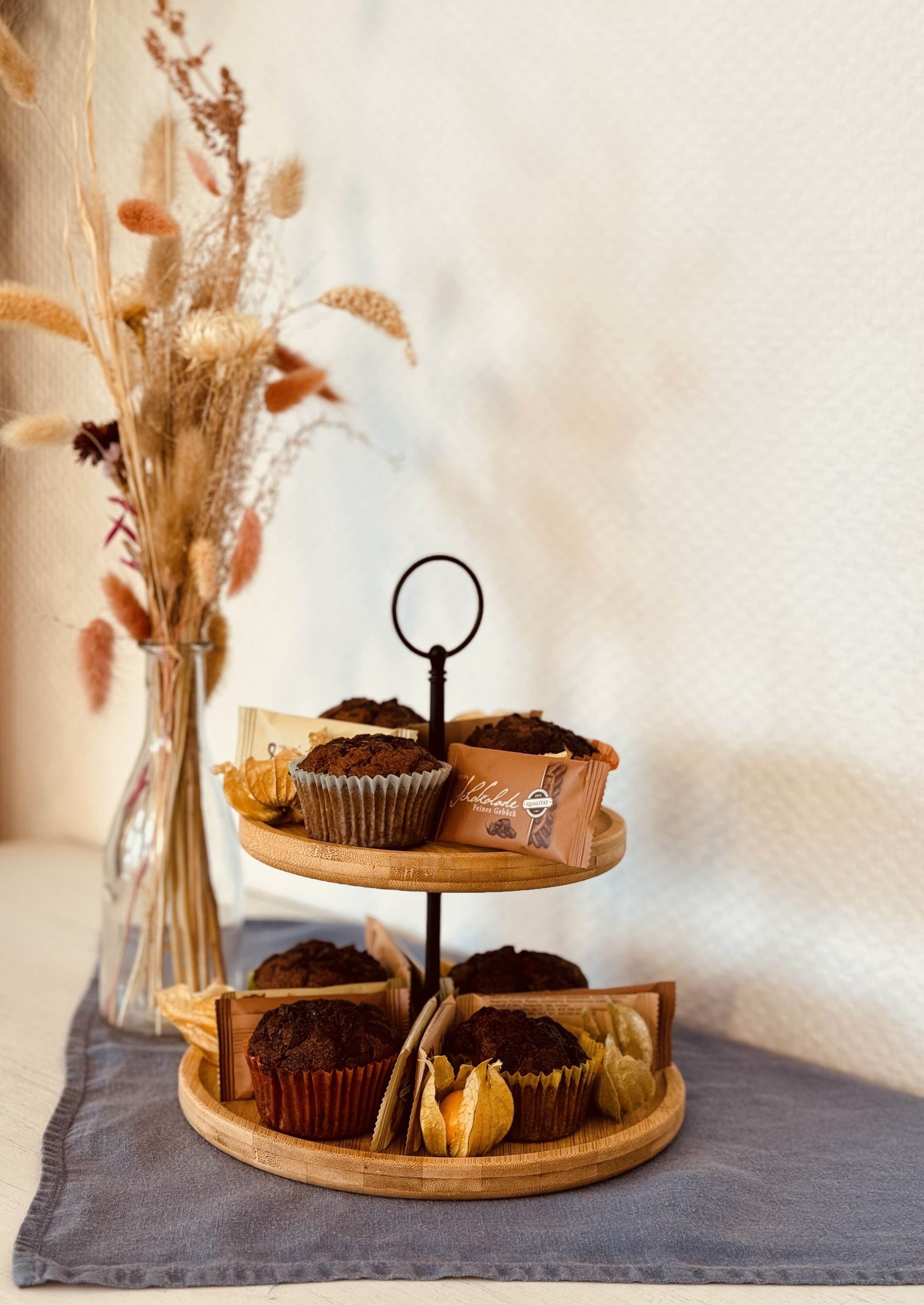 Un expositor de madera de tres niveles con varios productos horneados, un jarrón con flores secas y un mantel individual.