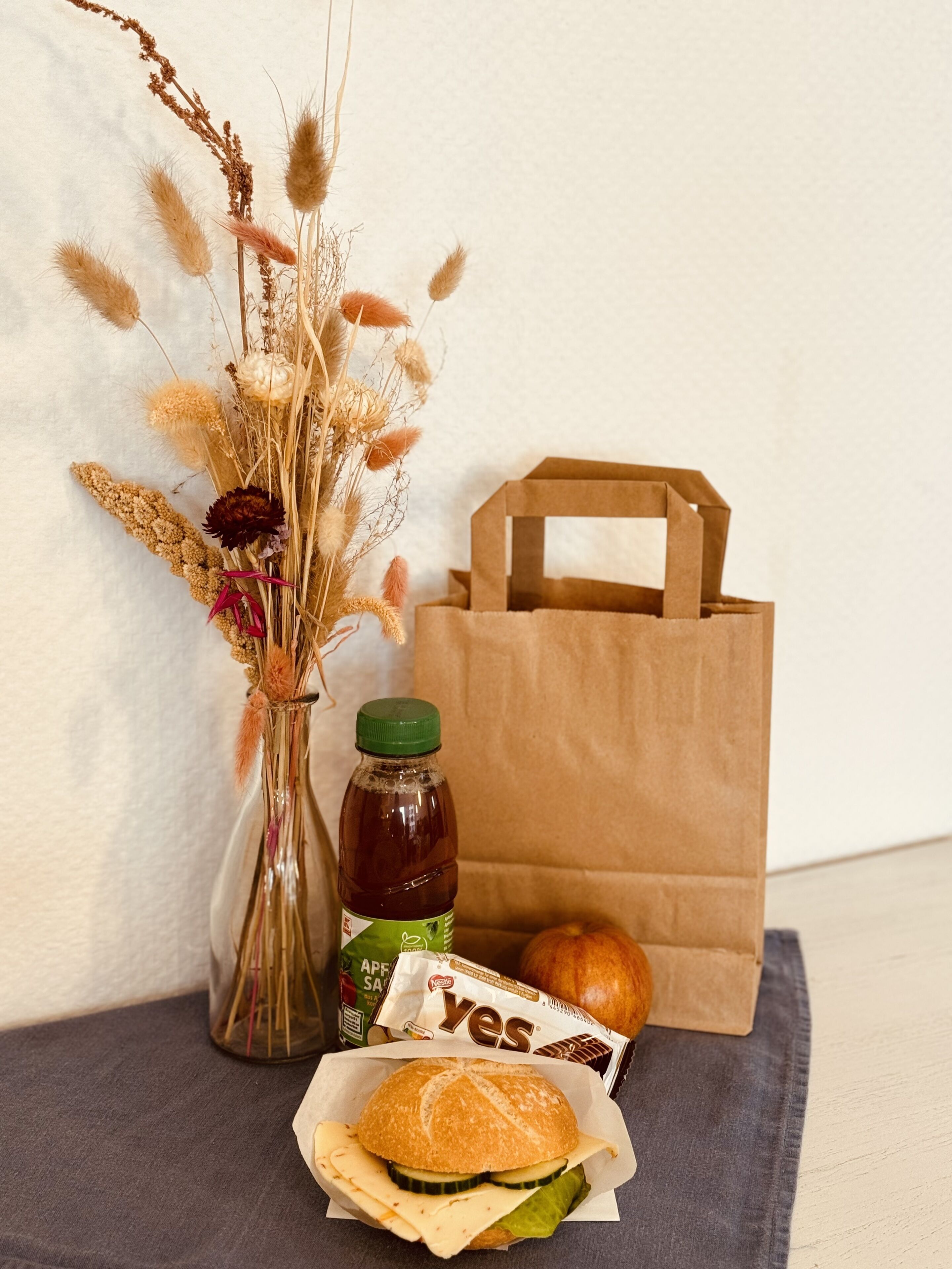 Een belegd broodje, een fles, een tas en gedroogde bloemen op een tafel.