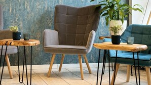 A modern lounge area with a textured blue wall, a tufted grey armchair, a green sofa, and two wooden tables with potted plants.