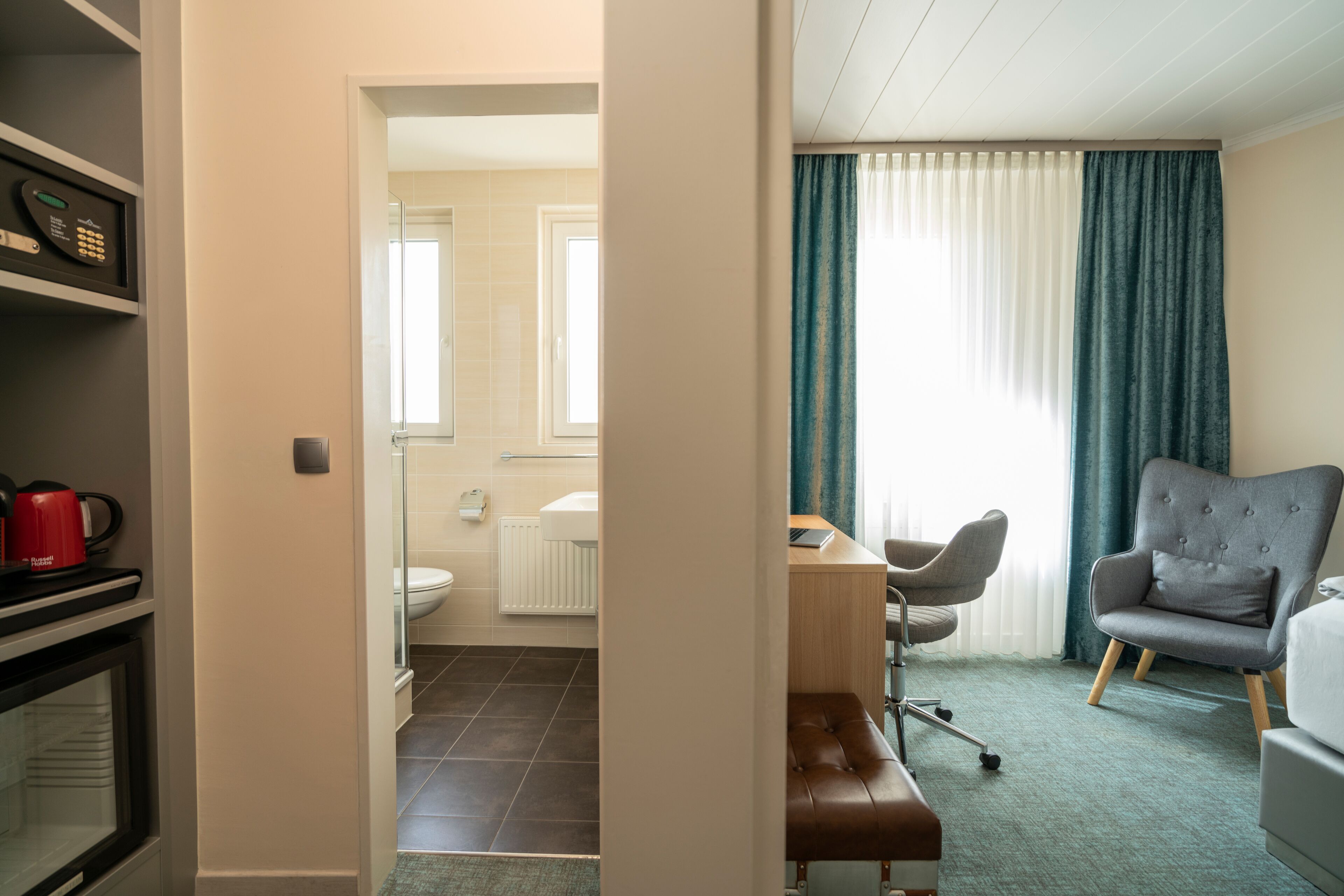 A hotel room with a kitchenette, a bathroom, a seating area with a chair and bench, and a large window with curtains.