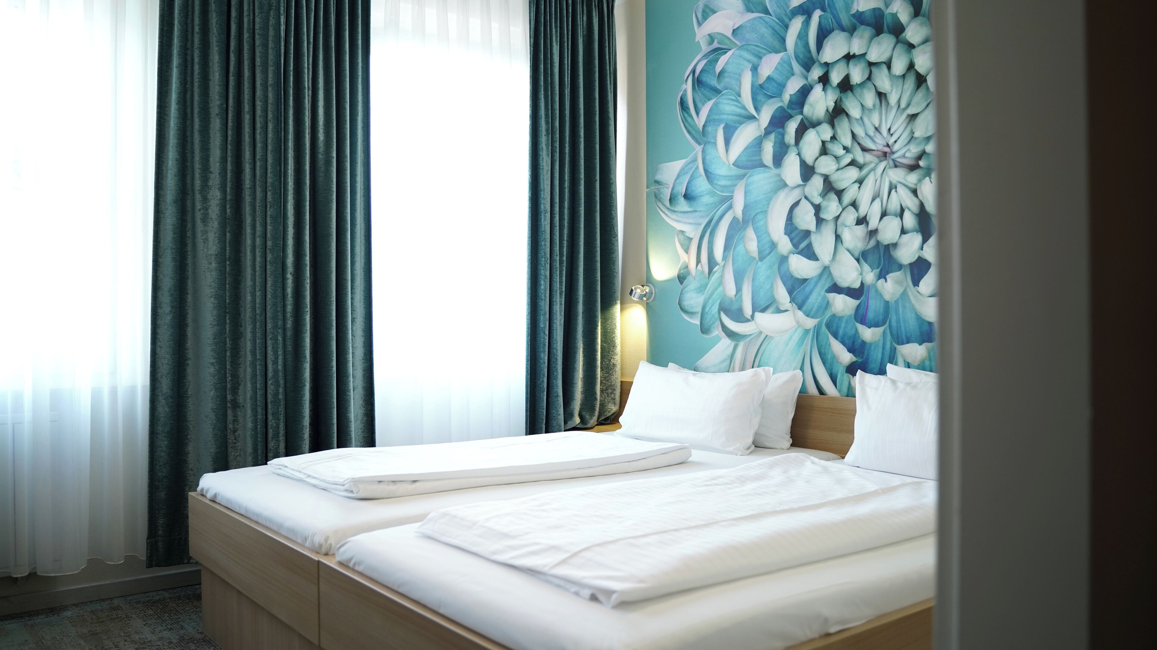 A hotel room with a bed, a large floral wall mural, and a window with curtains.