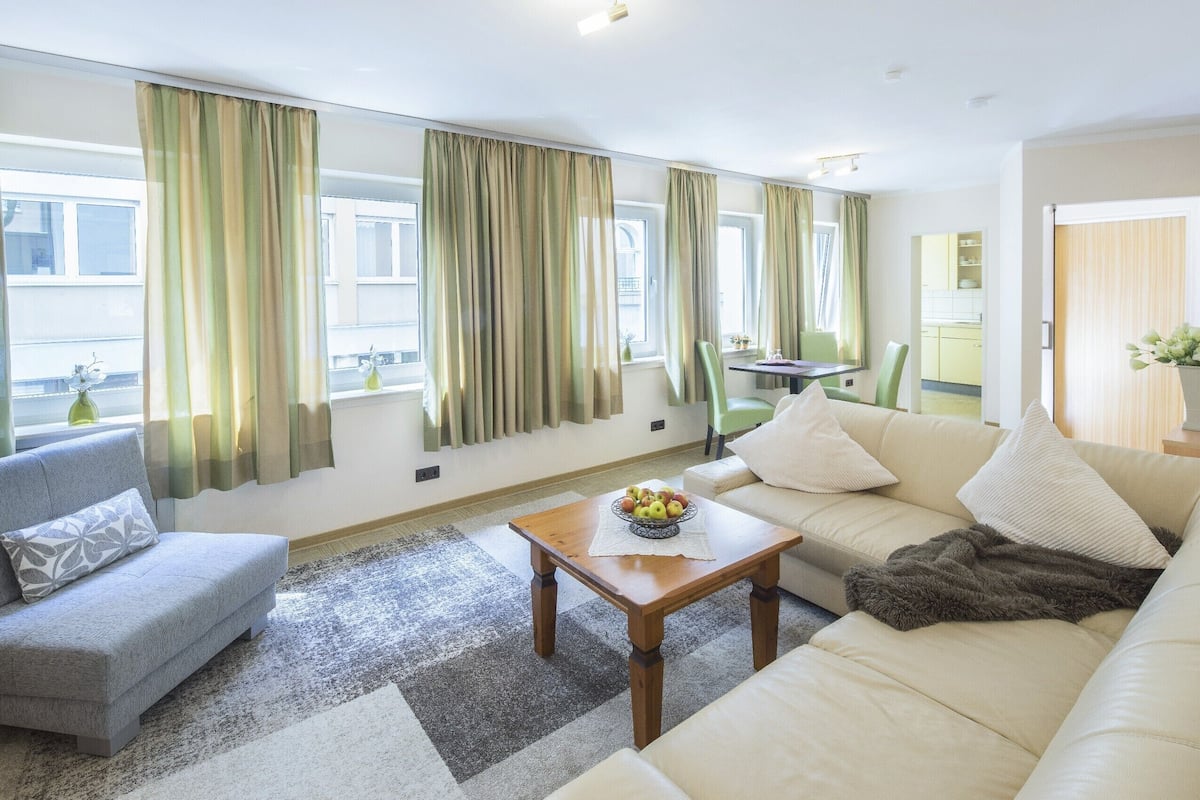 Ein modernes Wohnzimmer mit Sofa, Sessel und Couchtisch. Durch die Fenster ist ein Essbereich mit Tisch und Stühlen zu sehen.
