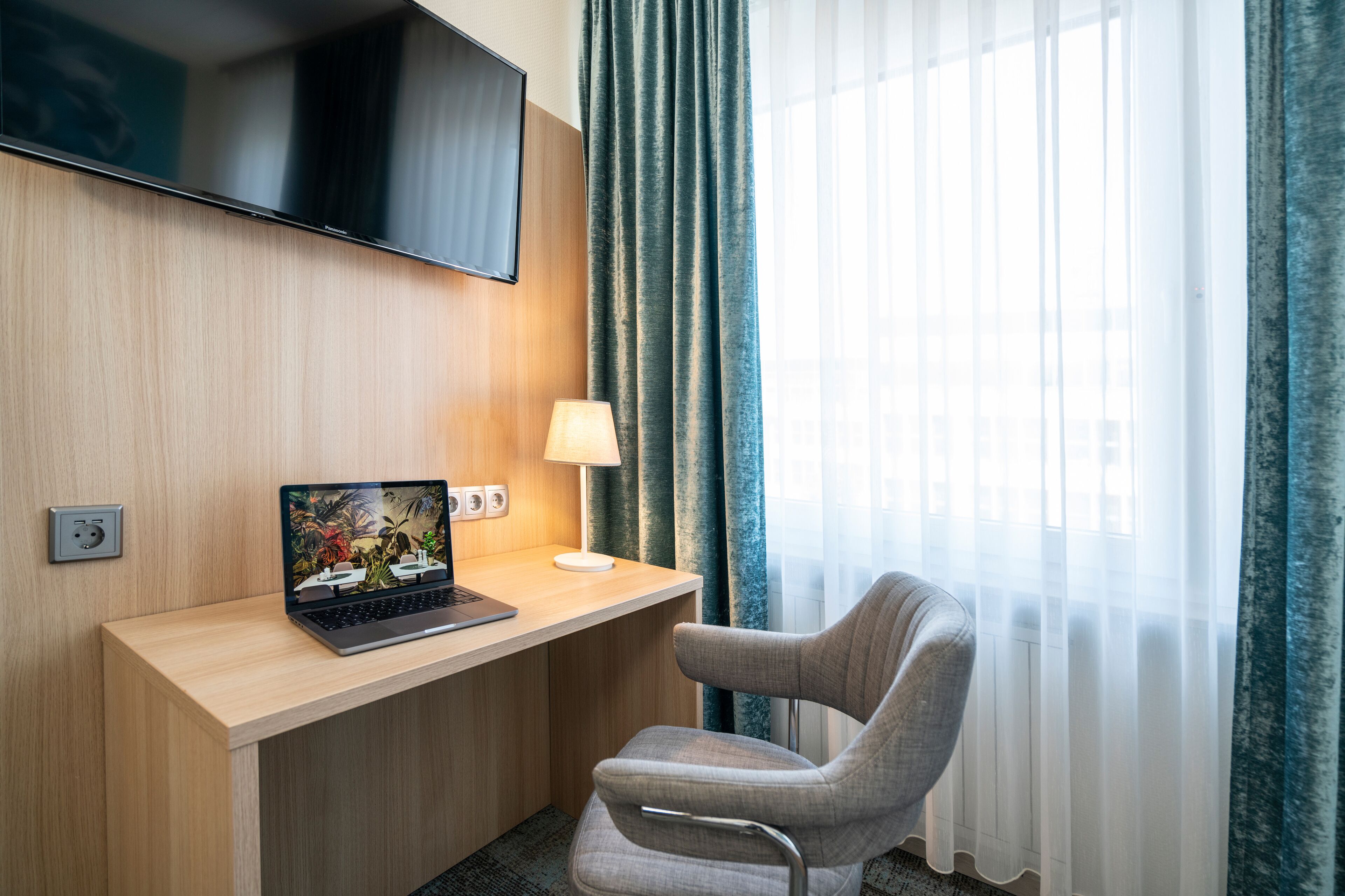 Ein Hotelzimmer mit einem Holztisch, einem Laptop, einer Lampe, einem an der Wand montierten Fernseher und einem Fenster mit Vorhängen.
