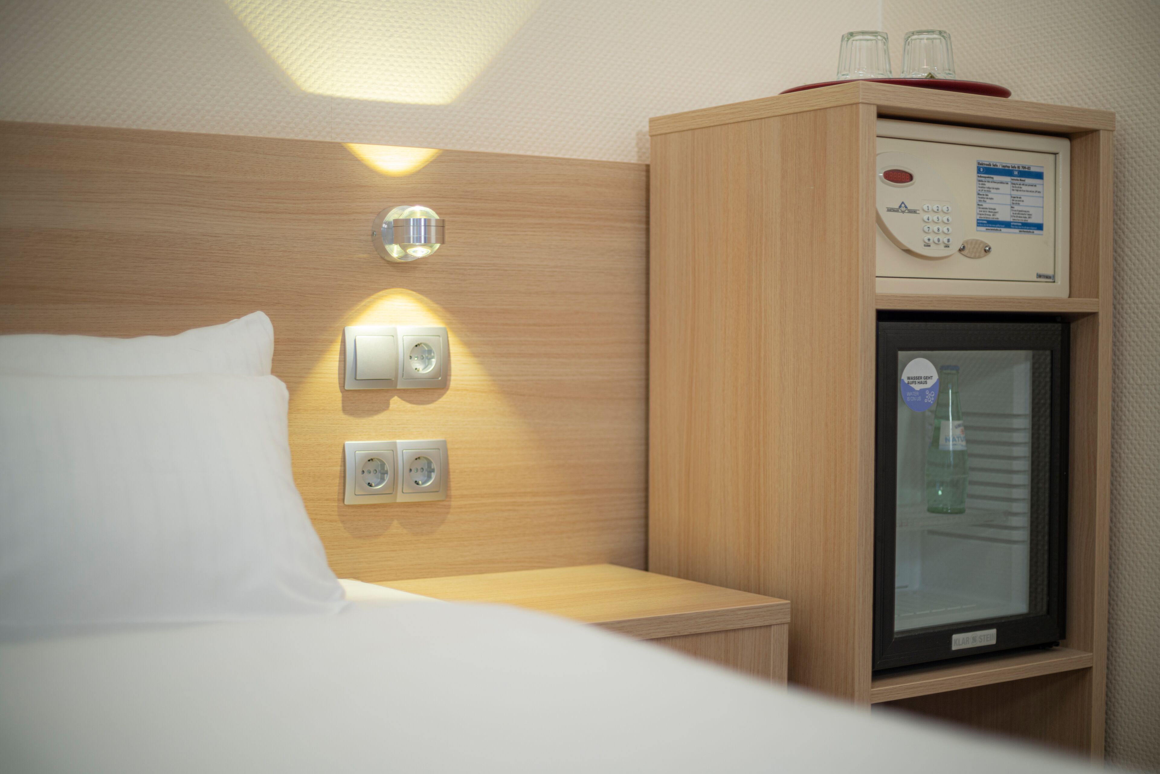 Ein Hotelzimmer mit Bett, Bedienelementen am Nachttisch, einer Minibar und einer Wandleuchte.