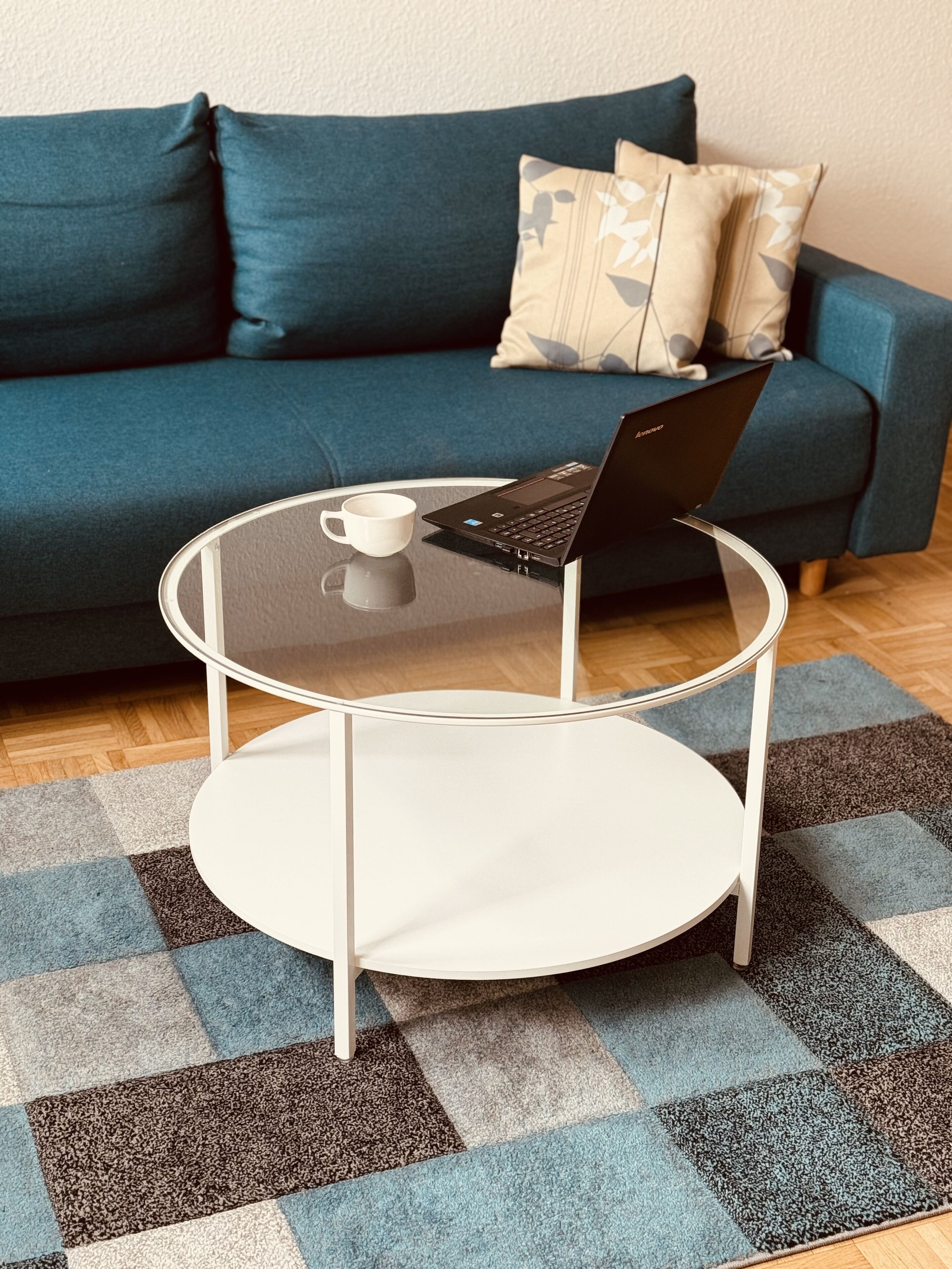 Ein Laptop steht auf einem Glastisch mit weißem Fuß, eine weiße Tasse und ein kariertes Teppich in Blau und Braun.