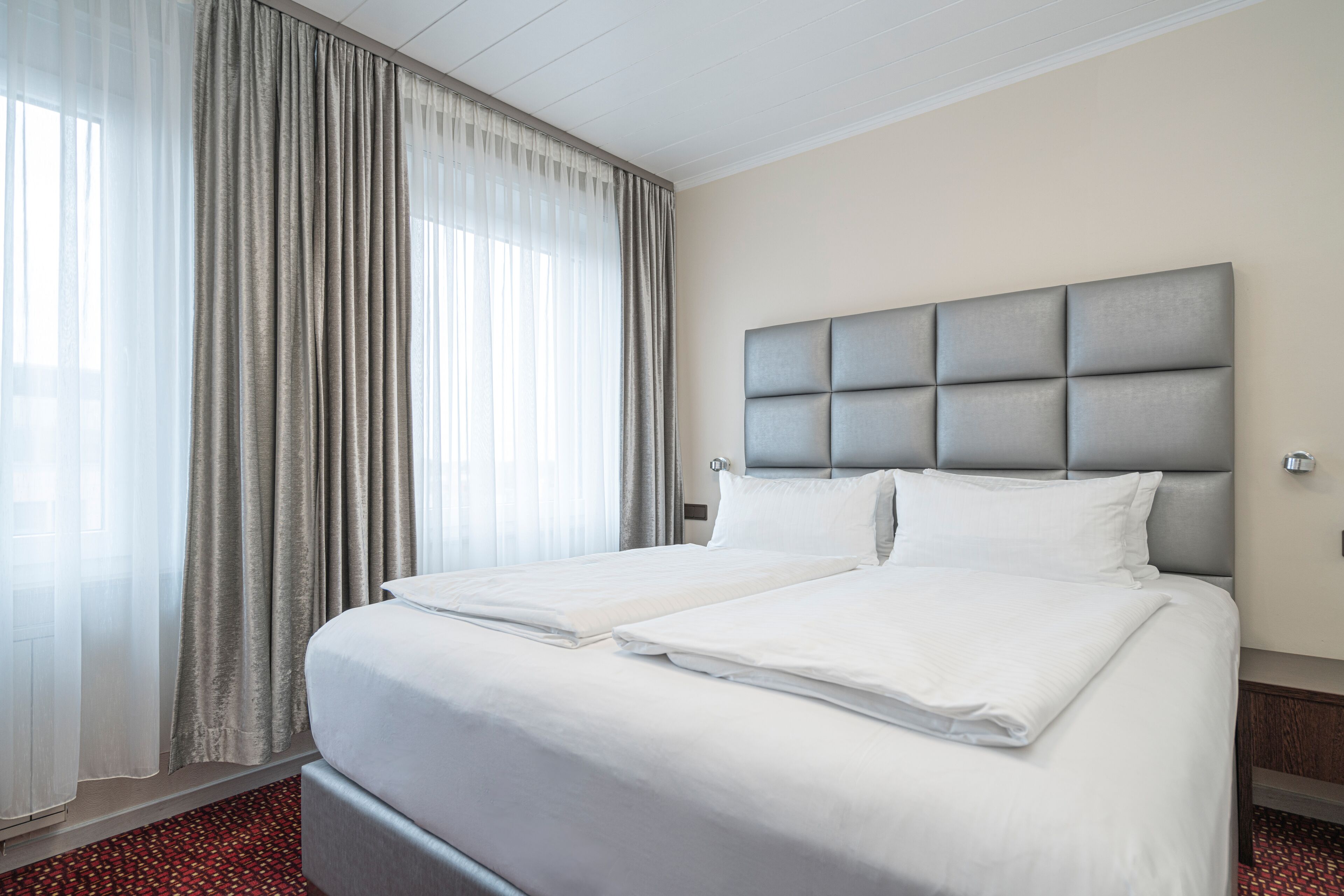 Una cama doble con cabecera tapizada de gris, ropa de cama blanca y alfombra roja estampada.