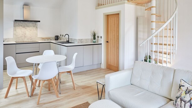 A modern kitchen with white cabinetry, a dining area with white chairs, and a staircase with wooden steps. - Host & Stay | Sunshine Cottage (Leyburn)